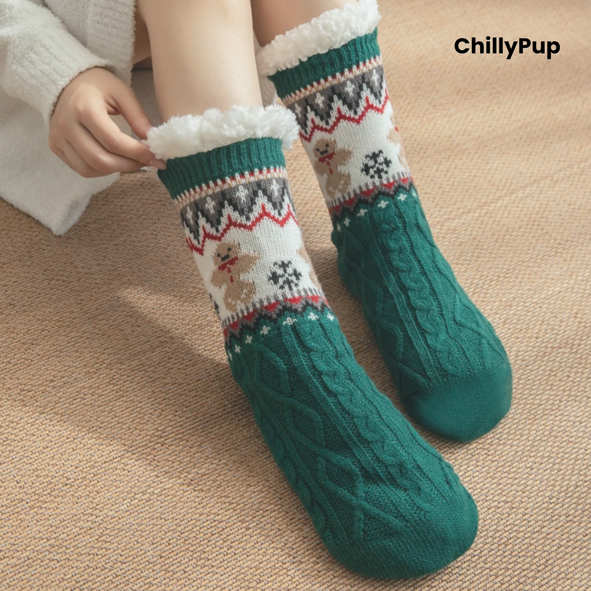 A person wearing festive Christmas-themed knit socks with a green cable-knit lower half, a cream patterned upper section with gingerbread motifs, and white faux-fur trim. The person is seated on a beige textured rug, wearing a white knit sweater.